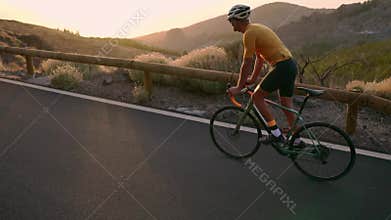 A professional cyclist in a helmet and sports equipment rides on a mountain highway at sunset in slow motion. Steadicam