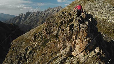 A young guy with a beard, a mountaineer in a cap and sunglasses, climbs a rocky ridge high in the mountains. Parkour in