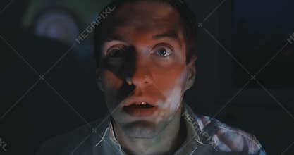 Close up of young man watching a horror video or film on TV or a computer monitor at home. He looks scared