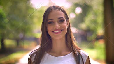 Nice lady is standing in park in daytime, watching at camera, smile, blurred backgrouns