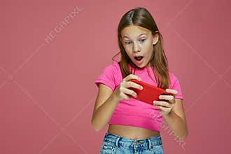 Amazed shocked child girl play game using mobile phone application on pink background. Children's addiction to gadgets