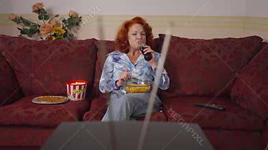 Relaxed happy retro woman eating chips drinking soda water sitting on couch watching vintage TV. Front view portrait of