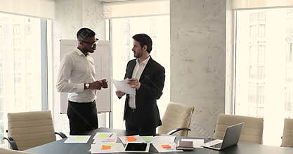 Two diverse businessmen consider documents, speaking, standing in boardroom