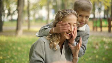 Close-up of relaxed beautiful slim woman sitting in autumn park as joyful boy running from background and closing her
