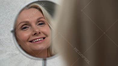 Happy 50s middle aged woman touching face skin looking in mirror reflection.