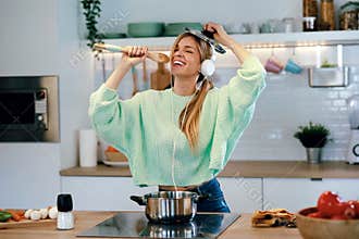 Beautiful young woman cooking while singing and listening music with headphones in the kitchen at home
