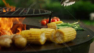Grilling vegetables cooking on fire outside. Man hands turning corn with forceps