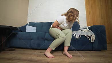 Woman in white T-shirt is sitting on blue sofa and holding her stomach with her hand in pain. Menstruation, pregnancy