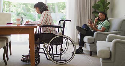 Happy biracial woman in wheelchair using laptop, with male partner talking on phone in living room