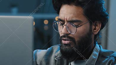 Close up Arabian Indian bearded 30s man freelancer user wear eyeglasses looking at computer screen at night at home has
