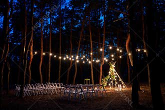 Festive string lights illumination on boho tipi arch decor on outdoor wedding ceremony venue in pine forest at night