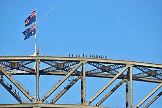 Adventurous daredevil climbing Sydney Harbour Bridge with Australia & NSW flags and clear blue sky in the background