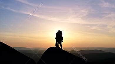 Slow motion of a man and a woman victoriously shake hands and embrace standing on a mountaintop at sunset.