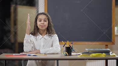 Portrait of intelligent beautiful African American schoolgirl raising hand smiling looking at camera. Middle shot of