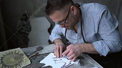 Bearded craftsman in apron making linocut with cutting tools