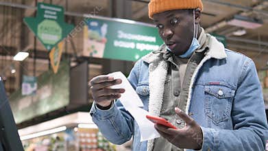 African-American man in a medical mask looks at a check at the checkout counter in a supermarket and is surprised and