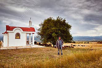 A shepherd in front of a little chapel and a herd of sheep