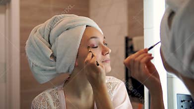 A woman with a white towel on her head applies eye shadow in front of the bathroom mirror. girl with brown eyes does a