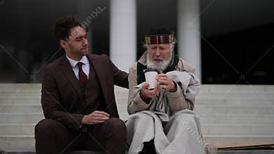 Kind businessman in suit giving takeaway coffee to homeless elderly man, showing compassion and generosity in urban