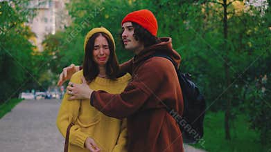 A young woman stands upset in the park. Her boyfriend hugs her, trying to comfort her, but despite his effort, she walks