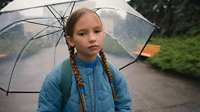 Tired little girl schoolgirl child looking at camera bored unhappy bad weather rain umbrella city park outdoors fatigue