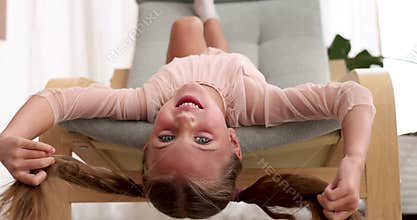 Smiling girl with ponytails hangs head down in armchair