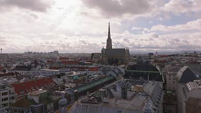 Forwards fly above buildings in city center. Tall church tower of Stephansdom against cloudy sky. Vienna, Austria