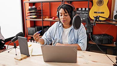 A middle-aged hispanic woman podcasting indoors with a laptop, microphone, headphones, and smartphone