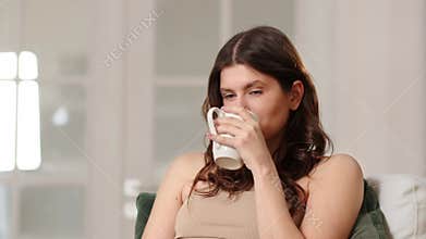 A beautiful brunette is sitting in an armchair with a cup of tea and thinking about something. A pleasant young woman