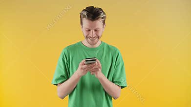 A smiling guy in a green T-shirt stands against a yellow wall with a phone in his hands. A young man is standing in