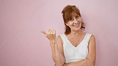 Cheerful middle age woman in a dress, standing by a pink wall, flashing a thumb up, pointing sideways with a glowing smile