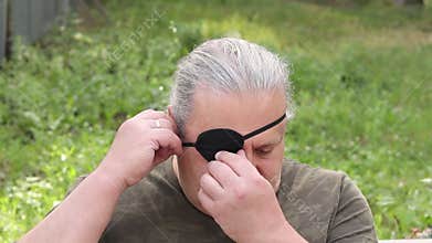 A blind, visually impaired man adjusts a black medical bandage on his eye while sitting on a park bench