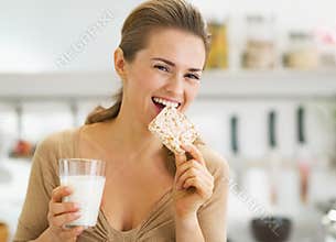 Happy young woman eating crisp bread with milk in kitchen