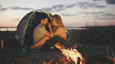 A serene and romantic scene unfolds as a couple enjoys a campfire at sunset, bonding and embracing in the wilderness