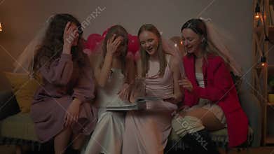 Group of young women friends and bride celebrating bachelorette party at home, bride and bridesmaids sit on sofa and