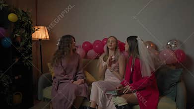 Group of young women friends and bride celebrating bachelorette party at home, bridesmaids sit on sofa holding photo