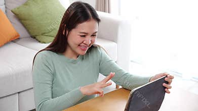 Young asian woman sitting on sofa using digital tablet computer for video call in living room at home.