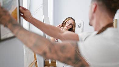 A tattooed man and spectacled woman collaborate in hanging a framed painting in a bright art studio