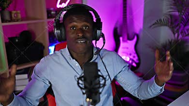 An african-american man podcasting with headphones and microphone in a colorful gaming room at night