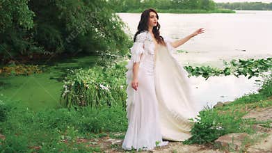 Fantasy woman sexy queen in long white dress stands fashion model posing by lake. Creative clothing, cloak cape with