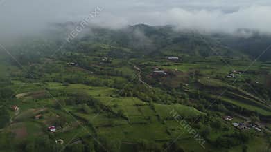 Flying above meadows and farm fields with cloudy sky and foggy background.