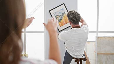 A man and woman collaborate in a bright art studio, hanging a colorful framed painting on a white wall