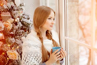 Beautiful young woman drinking tea at the Christmas tree. Beautiful girl celebrates Christmas with cup of cocoa.
