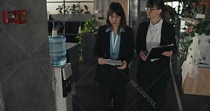 Two female colleagues walking down office corridor holding clipboards discussing work plans projects documents teamwork