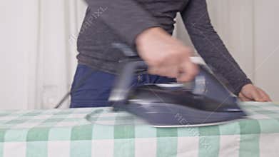 Man carefully irons a green and white checkered tablecloth on an ironing board, performing a household chore.
