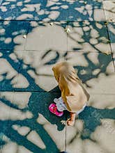 A young girl wearing a beige hijab is bending over, picking flowers on a paved walkway
