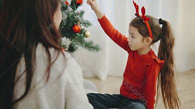 Mother and daughter sit together near a glowing Christmas tree, chatting and smiling in a cozy festive apartment, both wearing