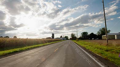 POV driving along rural American road between farmland fields with barn silo and dramatic clouds