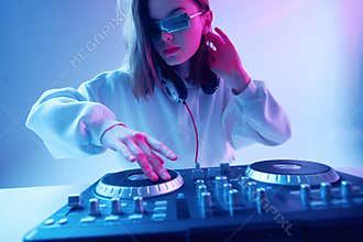 Cool young girl DJ mixes music on a mixing console and headphones, in stylish clothes, glasses on a neon background.