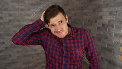 A young handsome man in plaid shirt scratches his head and looks at the camera on a gray brick background.